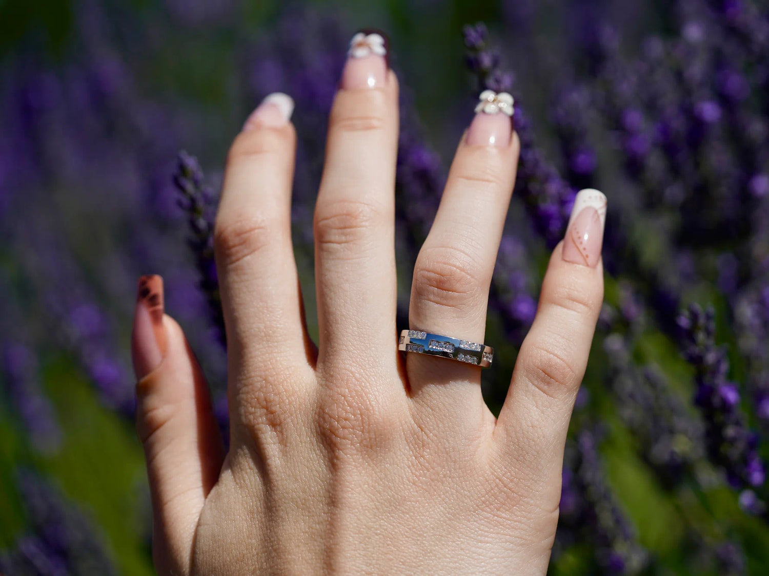 Nahaufnahme einer Hand mit liebevoll gestalteten Acrylnägeln und einem funkelnden, silbernen 3-reihigen Moissanite-Ring, vor einem blühenden Lavendelfeld