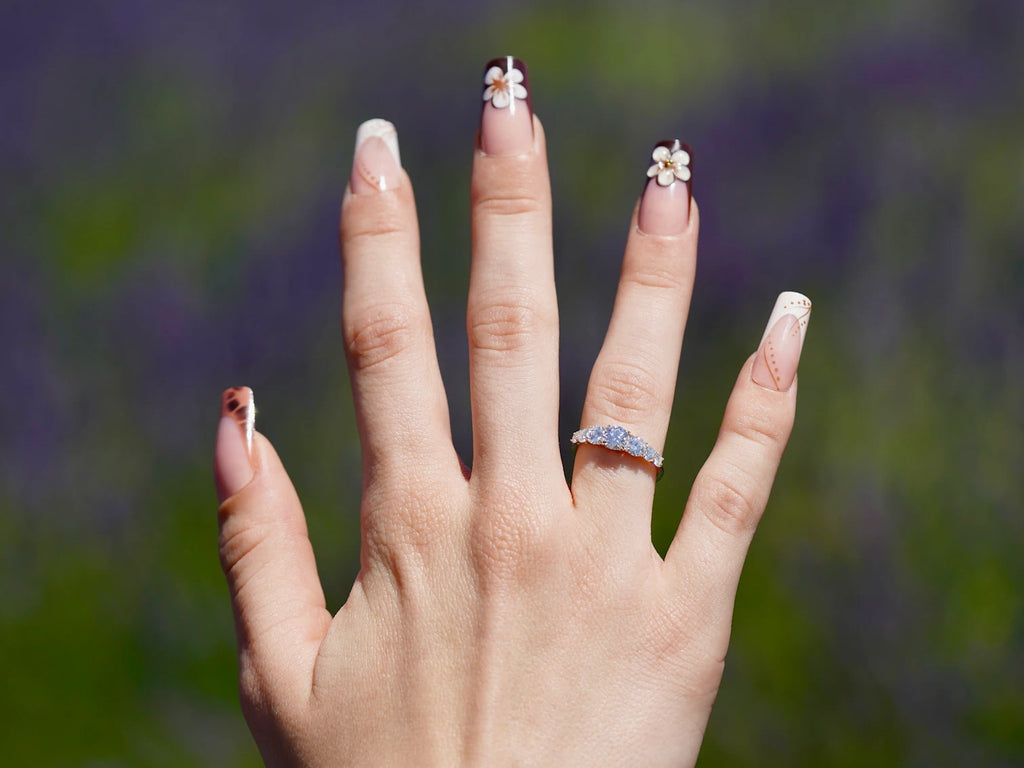 Nahaufnahme einer Hand mit liebevoll dekorierten Acrylnägeln, die einen funkelnden 5-Steine-Moissanite-Ring aus 925er Silber mit sanft abgestuftem Größenverlauf präsentiert, vor einem weichen, violetten Lavendel-Hintergrund.