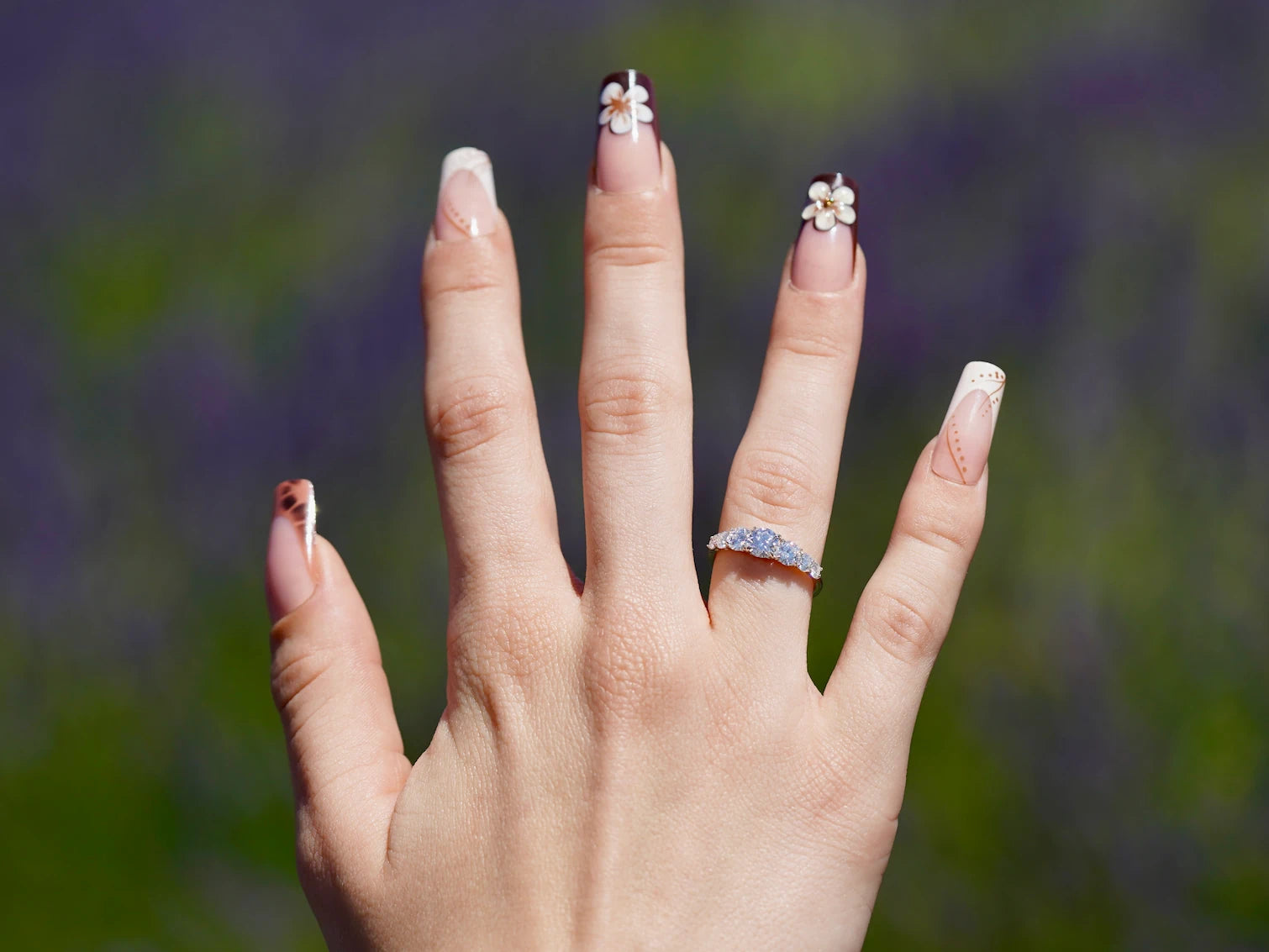 Nahaufnahme einer Hand mit liebevoll dekorierten Acrylnägeln, die einen funkelnden 5-Steine-Moissanite-Ring aus 925er Silber mit sanft abgestuftem Größenverlauf präsentiert, vor einem weichen, violetten Lavendel-Hintergrund.